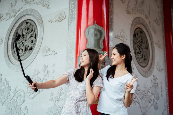 Dwie azjatyckie kobiety robiące selfie kijem akcji, pozujące ze zwycięską pozą dłoni. - Zdjęcie, obraz