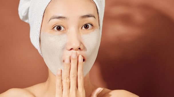 Gelukkig jong Aziatisch vrouw aanbrengen klei masker op haar gezicht over oranje achtergrond. Sluiten.. - Foto, afbeelding