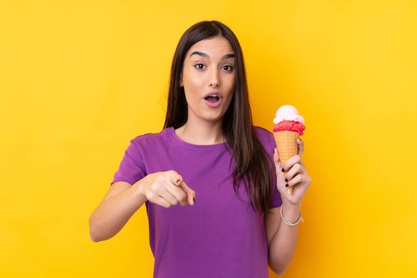Mladá brunetka žena s kornetovou zmrzlinou přes izolované žluté pozadí překvapen a ukazuje dopředu - Fotografie, Obrázek