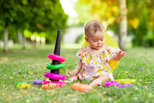 niña de 7 meses sentada en la hierba verde con un vestido y sombrero amarillo y jugando con una pirámide, desarrollo temprano de niños de hasta un año, caminando al aire libre
 - Foto, Imagen