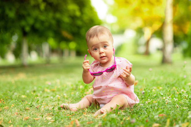 little baby girl 7 months old sitting on the green grass in a pink bodysuit and bright glasses, walking in the fresh air - Fotoğraf, Görsel