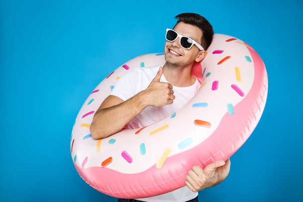 Young man with inflatable donut showing thumb up on blue background - Фото, зображення