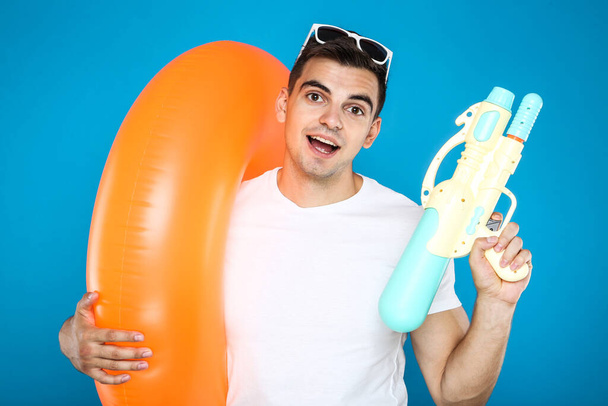 Young man with inflatable ring and water gun on blue background - Zdjęcie, obraz