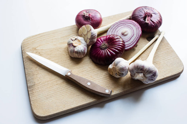 knife, red onion and garlic on wooden cutting board on white background - Photo, Image