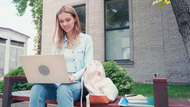 estudante sorrindo usando laptop perto de livro e cadernos no banco  - Filmagem, Vídeo