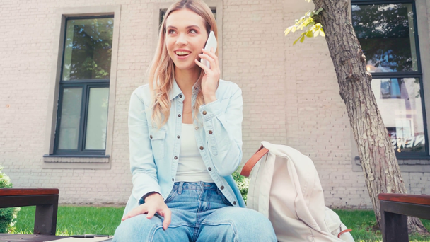 Estudante sorrindo conversando no smartphone perto da mochila no banco na rua urbana
  - Filmagem, Vídeo