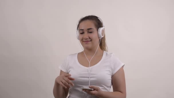Beyaz tişörtlü genç bir kadın kulaklıkla müzik dinlemek için telefon kullanıyor. - Video, Çekim