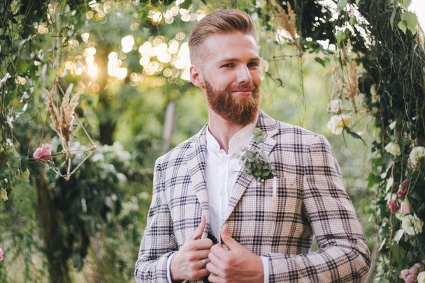 Portrait de mariage d'homme avec barbe rouge et coiffure hipster dans des vêtements vintage élégants. Mariage barbu posant en veste à carreaux à l'extérieur dans le jardin. Modèle masculin style rustique le jour du mariage dans la nature. - Photo, image