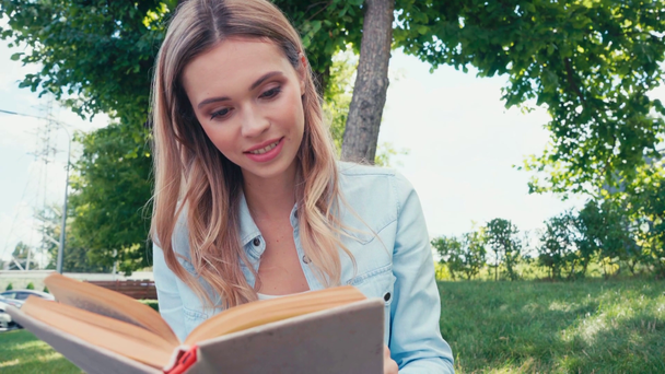 Όμορφη φοιτήτρια χαμογελώντας, ενώ ανάγνωση του βιβλίου στο πάρκο  - Πλάνα, βίντεο