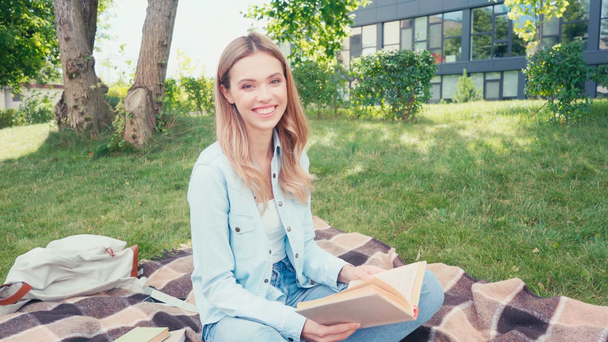 Smiling student waving hand at camera while reading book on plaid in park - Footage, Video