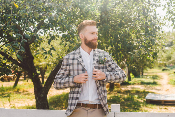 Wedding portrait of man with red beard and hipster hairstyle in stylish vintage clothing. Bearded groom posing in plaid jacket outdoors in the garden. Male model rustic style on wedding day in nature. - Фото, изображение