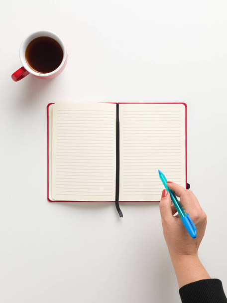 Vista superior de un cuaderno rojo en blanco abierto en el centro, una taza de café rojo y una mano femenina sosteniendo una pluma azul en fondo blanco, con espacio para texto - Foto, Imagen