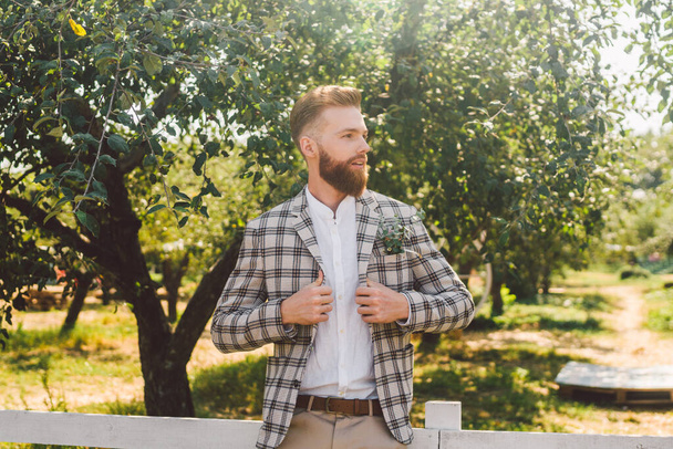 Portrait of Caucasian stylish bearded groom in vintage suit posing outdoors in sunny weather. Handsome man with beard in plaid jacket. Male model at wedding in rustic style. Groom in checkered jacket. - Foto, immagini