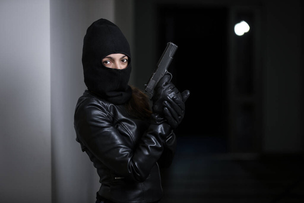A tolvaj betört a lakásba. Fekete kabátos és fekete maszkos nő rabolta ki a házat, fekete pisztollyal a kezében. Betörő maszkban. Tolvaj maszkban próbál betörni mások házába. - Fotó, kép