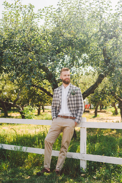 Wedding portrait of man with red beard and hipster hairstyle in stylish vintage clothing. Bearded groom posing in plaid jacket outdoors in the garden. Male model rustic style on wedding day in nature. - Photo, Image