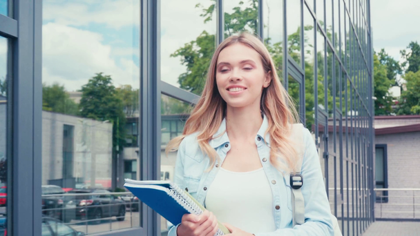 Χαμογελώντας φοιτητής με βιβλίο και σημειωματάρια με τα πόδια κοντά στο κτίριο σε αστικό δρόμο  - Πλάνα, βίντεο