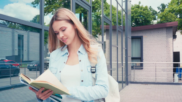 Улыбающийся студент читает книгу во время прогулки по городской улице  - Кадры, видео