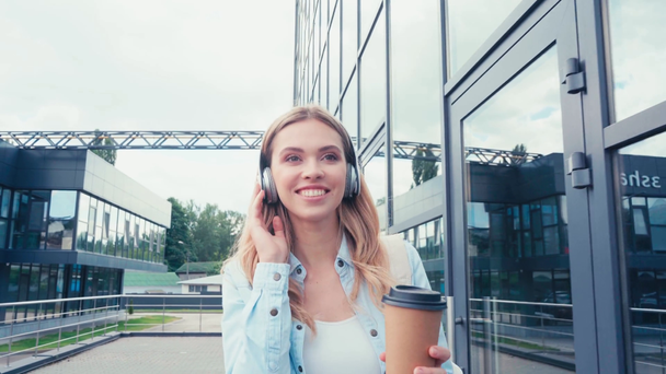 szczęśliwy student w słuchawki trzymając kawę iść podczas spaceru po miejskiej ulicy - Materiał filmowy, wideo