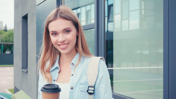 Student pijący kawę na wynos i uśmiechający się w pobliżu budynku na ulicy miejskiej - Materiał filmowy, wideo