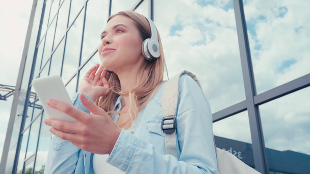 Estudante alegre em fones de ouvido segurando smartphone perto de edifício na rua urbana
 - Filmagem, Vídeo