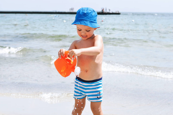 Menino bonito na praia em calções de banho listrados azuis corre nas ondas com uma lata de rega de plástico
 - Foto, Imagem