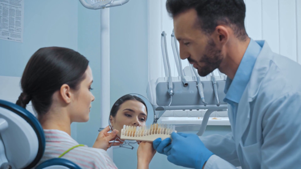 Dentysta i pacjent z lustrem patrzący na paletę zębów w klinice  - Materiał filmowy, wideo