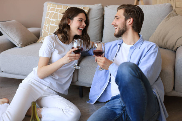Happy couple sitting, relaxing on floor in living room, drinking red wine. Smiling young husband and wife rest at home enjoy romantic date on family weekend together. - Zdjęcie, obraz