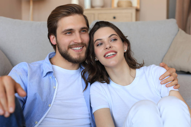 Young loving couple relaxing on floor near sofa together. Woman and man embrace enjoy company of each other sitting on couch holding hands having romantic date - Foto, Imagem