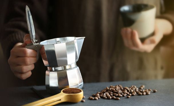 Vrouw in handen houden traditionele moka pot koffiezetapparaat klaar om verse koffie te gieten in de kleine kop. Koffiebonen en gemalen koffie op de achtergrond. - Foto, afbeelding