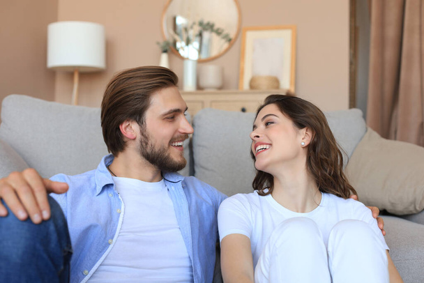 Young loving couple relaxing on floor near sofa together. Woman and man embrace enjoy company of each other sitting on couch holding hands having romantic date - Foto, afbeelding
