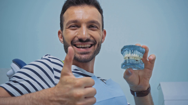 Улыбающийся пациент с моделью зубов и показывающий большой палец в клинике - Кадры, видео