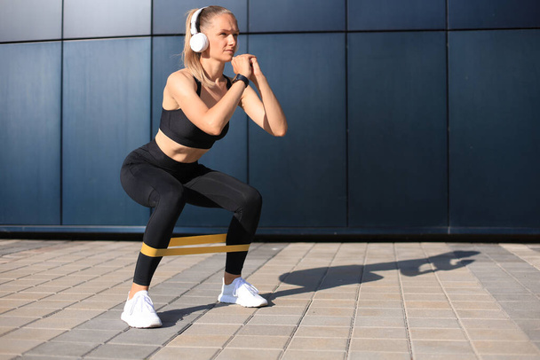 Sporty woman doing squats with fitness gum expander outdoors - 写真・画像