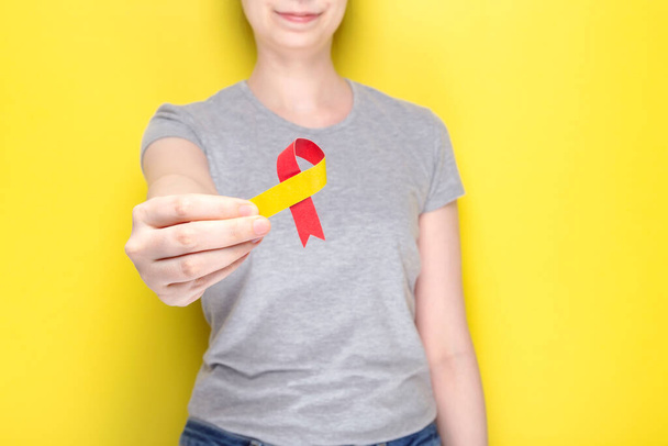 World Hepatitis Day concept. Girl in gray T-shirt holds in her hand awareness symbol red-yellow ribbon. Yellow background - 写真・画像