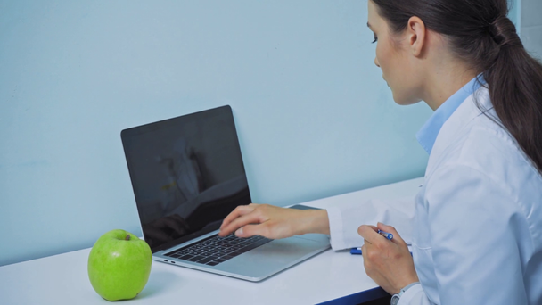 Tandarts met behulp van laptop en schrijven op klembord in de buurt van appel op tafel  - Video