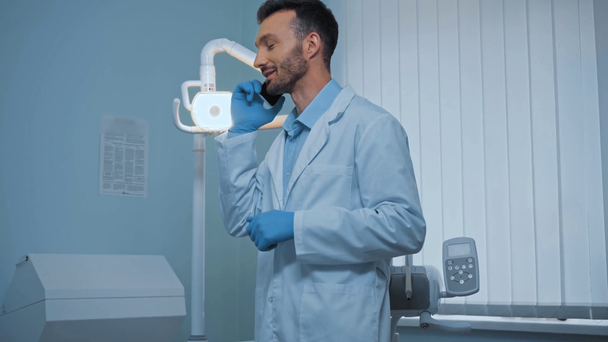 Hezký zubař mluví na smartphonu na klinice - Záběry, video