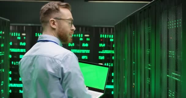 緑の画面のノートパソコンとサーバーの間を歩くメガネで白人の若い男のリアとビッグデータプロセッサの仕事をチェックします。クロマキー。バックビュー。データストレージにおける男性分析. - 映像、動画