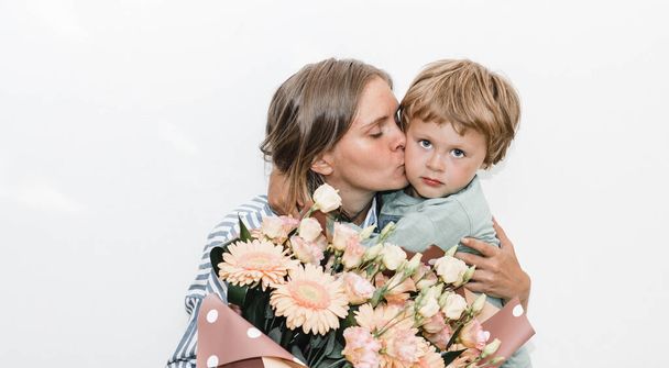 Ο γιος και η νεαρή μητέρα αγκαλιάζονται και φιλιούνται. το παιδί συνεχάρη τη μητέρα του για τα γενέθλιά της και παρουσίασε μπουκέτο  - Φωτογραφία, εικόνα