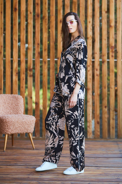 Young stylish sexy woman in pajamas standing on terrace in tropical hotel, palm trees background, long black hair, sunglasses, earrings, sunglasses. - Photo, Image