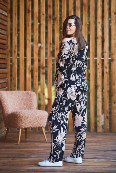 Young stylish sexy woman in pajamas standing on terrace in tropical hotel, palm trees background, long black hair, sunglasses, earrings, sunglasses. - Valokuva, kuva
