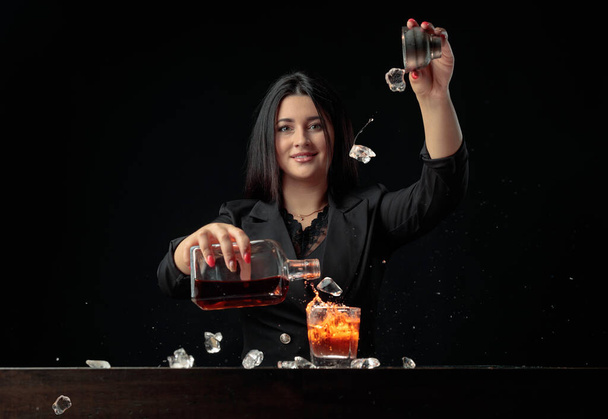 Attractive bartender girl pours an alcoholic drink into a glass and throws pieces of ice. Beautiful brunette in black prepare cocktail. - Photo, Image