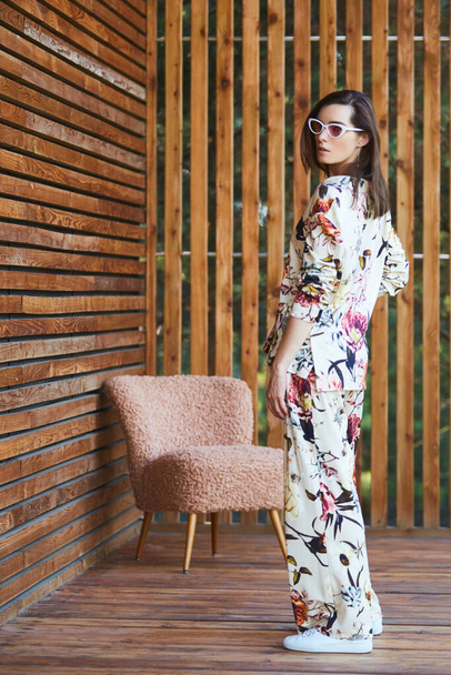 Young stylish sexy woman in pajamas standing on terrace in tropical hotel, palm trees background, long black hair, sunglasses, earrings, sunglasses. - Φωτογραφία, εικόνα