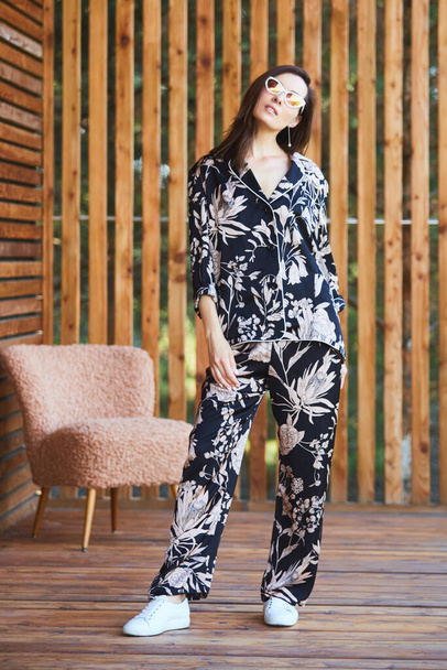 Young stylish sexy woman in pajamas standing on terrace in tropical hotel, palm trees background, long black hair, sunglasses, earrings, sunglasses. - Photo, Image