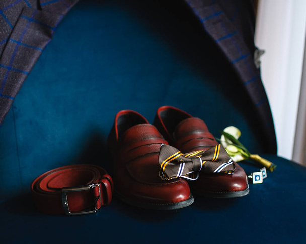 Elegantes accesorios de novio, detalles del atuendo del novio: zapatos marrones, cinturón y pajarita en una silla azul profundo
 - Foto, Imagen