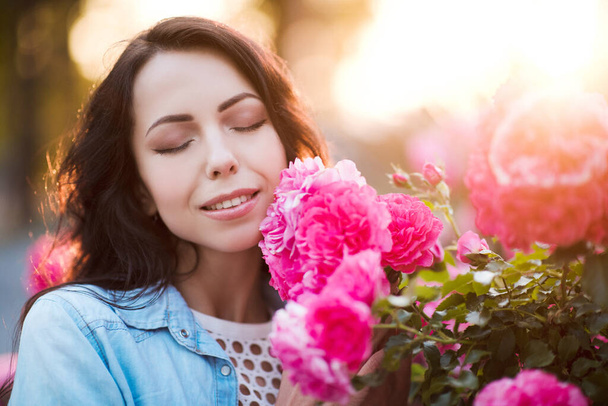 Mujer sonriente de 24-25 años oliendo rosas al aire libre de cerca. Temporada de verano. 20s
.  - Foto, Imagen