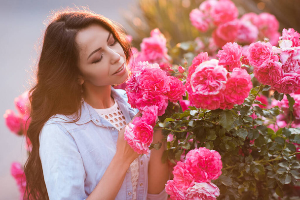 Uśmiechnięta piękna kobieta 20-24 lat pachnąca różowymi różami w ogrodzie na zewnątrz zbliżenie. Sezon letni. Romantyzm. - Zdjęcie, obraz