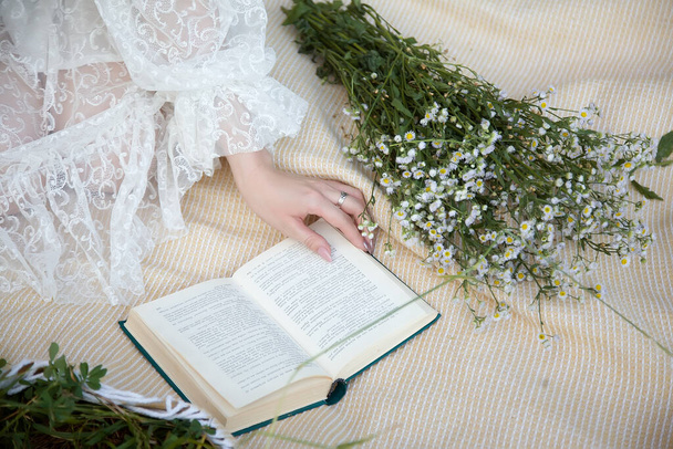 On a bedspread in the grass, a book and a bouquet of wild flowers, a girl in a lace dress, her hand turns the pages. White lace vintage dress. Picnic - Фото, изображение