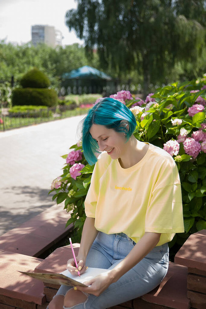 Une femme artiste aux cheveux bleus s'assoit dans un parc et dessine avec un carnet de croquis près d'une hortensia en fleurs. Peinture femme plus âgée en plein air - Photo, image