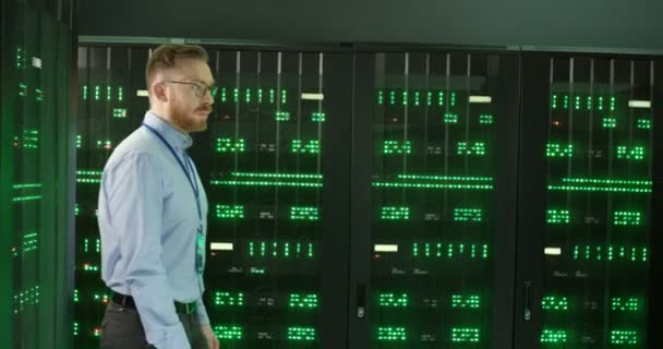 Caucasian handsome young man in glasses coming in and walking among servers with big data information. Green working processors in database storage. Male worker of digital telecommunication. - Footage, Video