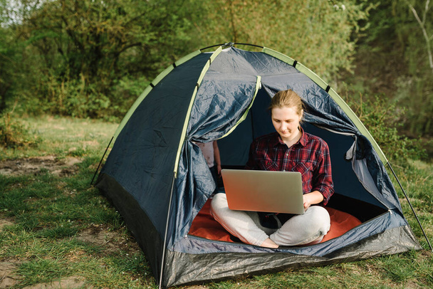 Woman working on laptop in tent in nature. Young freelancer sitting in camp. Relaxing in camping site in forest, meadow. Remote work, outdoor activity in summer. Happy girl relaxing, work on vacation. - Foto, afbeelding