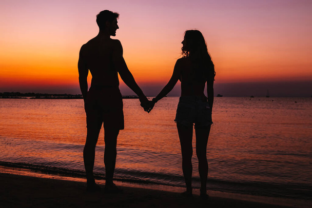 влюбленная пара на пляже на закате - силуэт молодых влюбленных при закате солнца - Фото, изображение